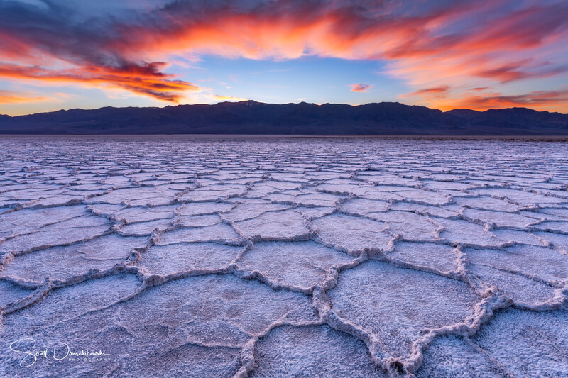 Salt flat with hexagonal crystalline patterns under dramatic sunrise sky with orange and blue clouds.