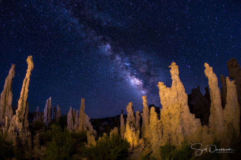 Milky Way galaxy arcing across night sky above illuminated rock formations in desert landscape.