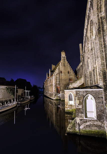 Medieval church illuminated at night beside a calm waterway, with Gothic architecture visible across the water.