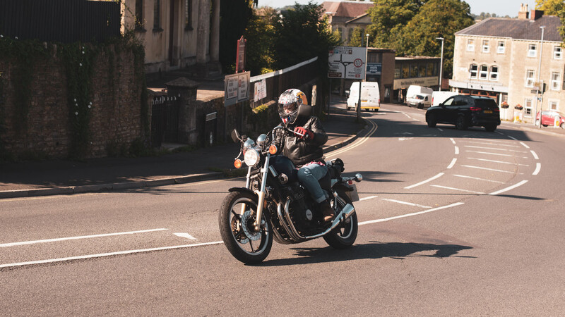 Motorcyclist riding a vintage-style cruiser through a town street intersection on a sunny day.