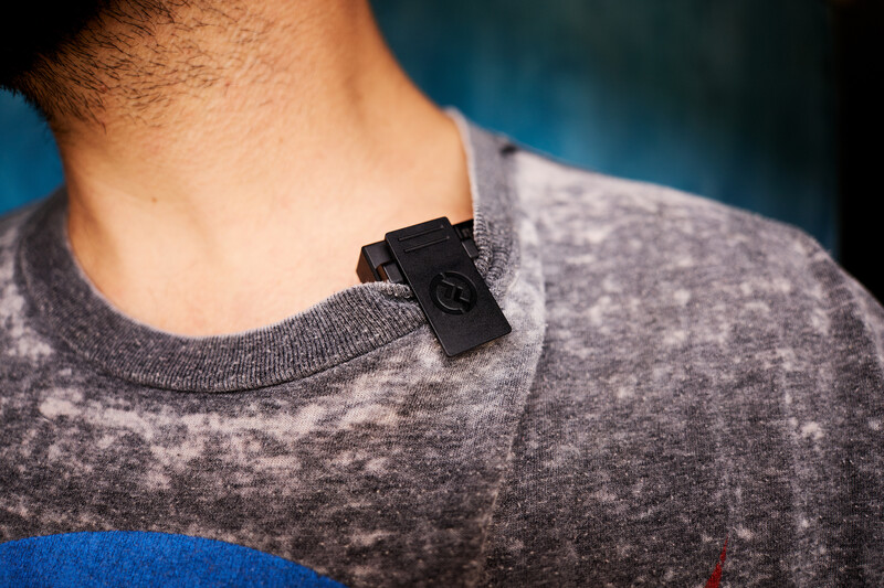 Small black microphone clip mounted on gray t-shirt collar against blurred blue background.