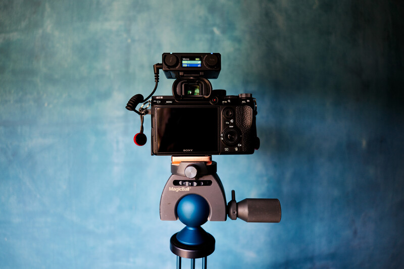 Professional video camera rig mounted on tripod with external monitor and microphone against textured blue backdrop.