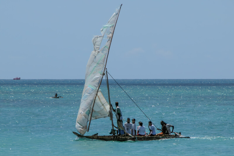 Traditional sailing vessel with white sail and multiple passengers on turquoise ocean water.
