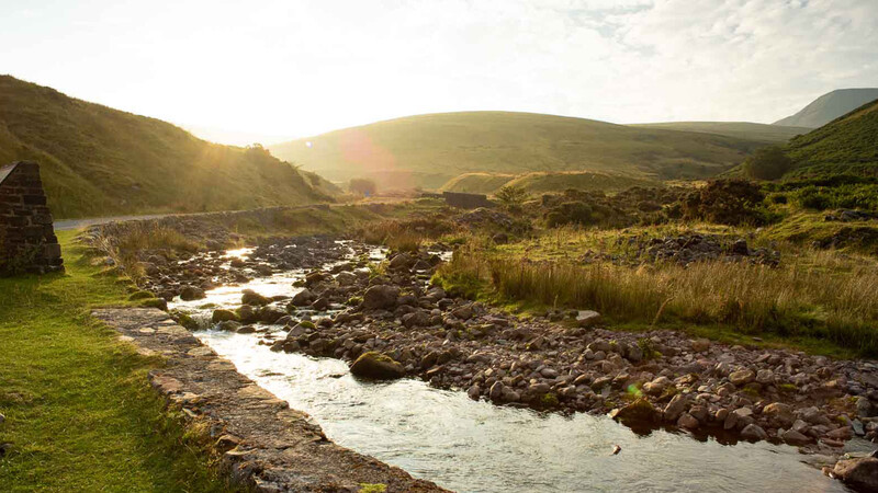 Mountain valley landscape with winding stream, golden hour light, and rolling green hills.