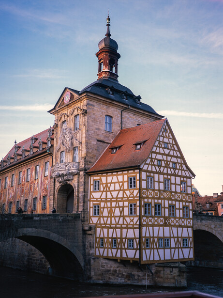 Half-timbered building with red tile roof and church tower reflected in water below.