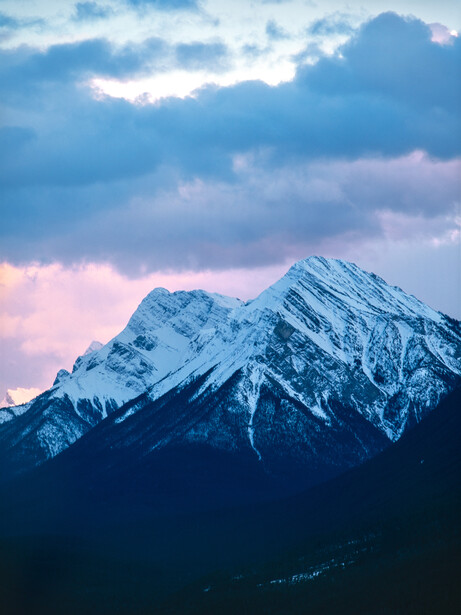 Snow-covered mountain peak lit by pink and blue alpenglow at dusk beneath dramatic clouds.