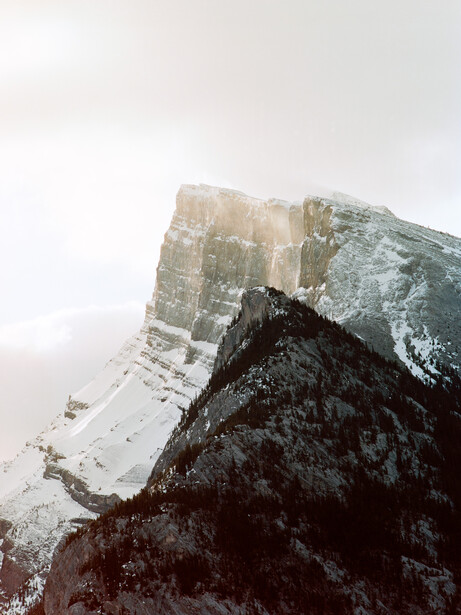 Snow-covered mountain peaks with dark forested slope in foreground under overcast sky.