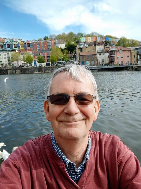 Man wearing sunglasses and maroon cardigan smiling at camera with waterfront buildings behind him.