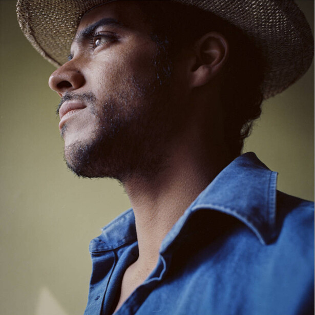 Man in profile wearing a straw cowboy hat and blue denim shirt against a neutral background.