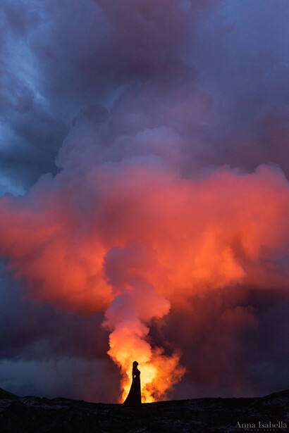 Silhouetted figure standing before an active volcanic eruption with dramatic orange lava and dark storm clouds.