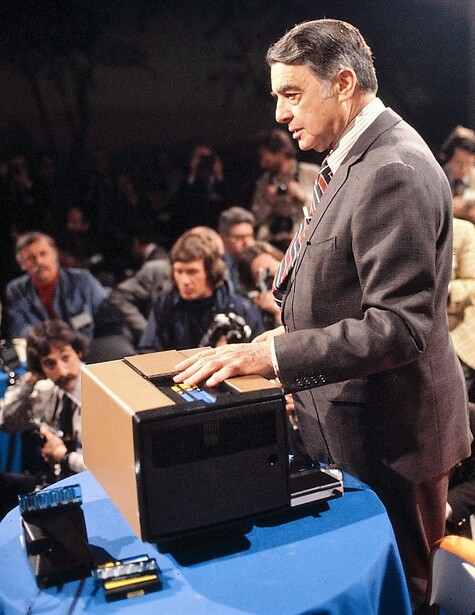 Man in blazer speaking at podium with blue front panel during presentation to seated audience.