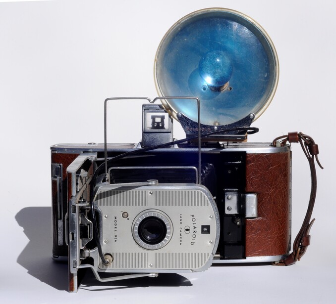 Vintage instant camera with brown leather case and blue flash attachment mounted on top.