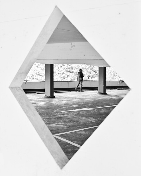 Black and white photograph of a person standing on a concrete platform beneath a covered structure with support columns.