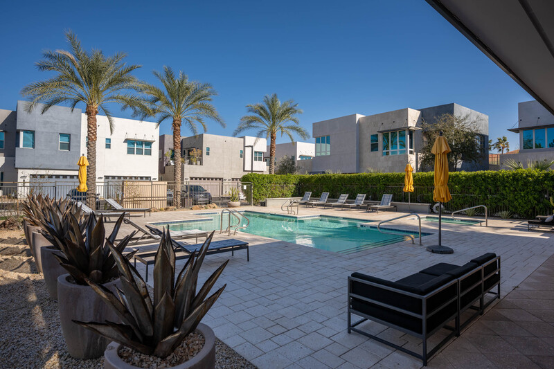 Community pool area with loungers, surrounded by modern residential buildings and palm trees under clear sky.