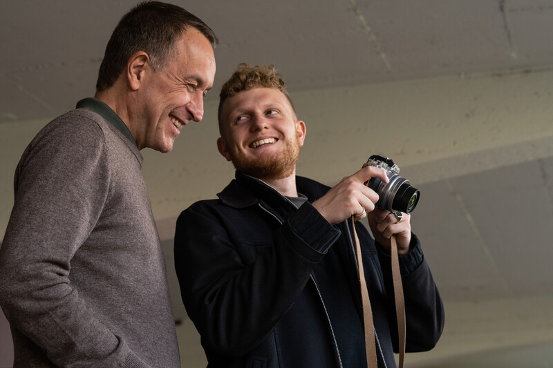 Two men smiling while holding a mirrorless camera during an indoor discussion or demonstration.
