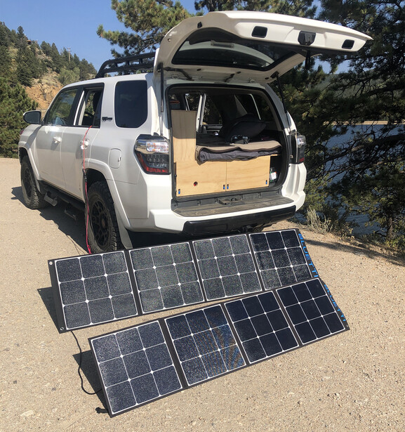White SUV with open rear hatch showing wooden interior storage, with portable solar panels deployed on ground in front.