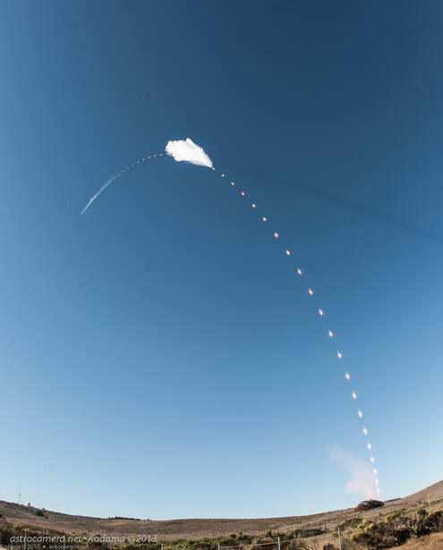 Rocket launch trail arcing across clear blue sky above desert terrain.