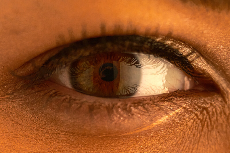 Close-up of human eye with warm golden-brown skin tone, hazel iris with red undertones, and detailed eyelashes.