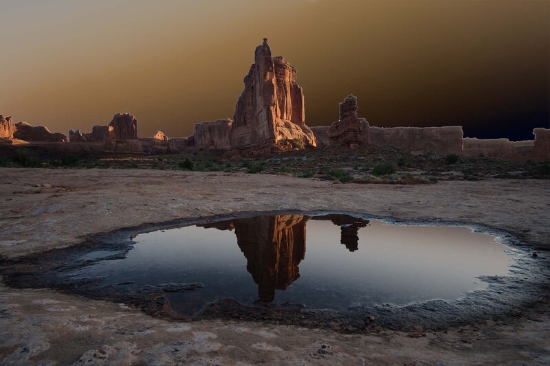 Tall rock formation reflected in still water at sunset in desert landscape.