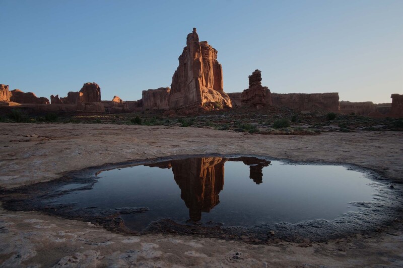 Towering red rock formation reflected in still water pool at desert landscape during golden hour.