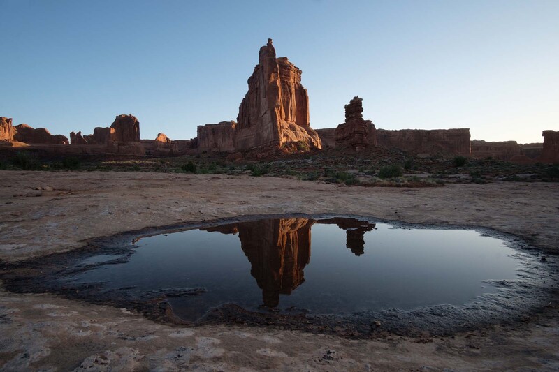 Tall red rock formation reflected in still water pool at desert landscape during golden hour.