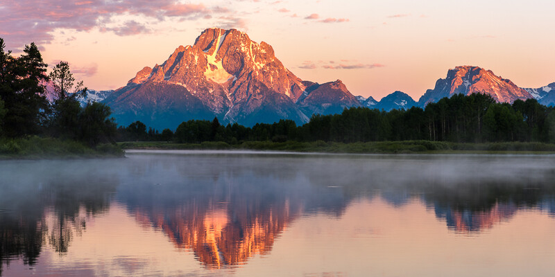 Mountain peak reflected in calm lake at sunrise with forested shoreline and mist.