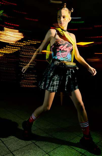 Young woman in graphic tank top and plaid skirt posed against neon-lit background.