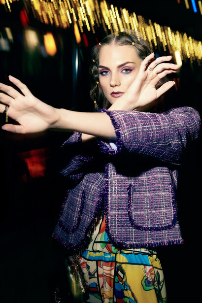 Woman in purple tweed jacket with colorful patterned belt posing with arms extended against warm-toned background.