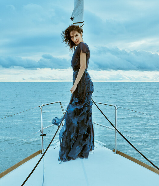 Woman standing on boat bow wearing flowing blue patterned dress against calm ocean and sky.