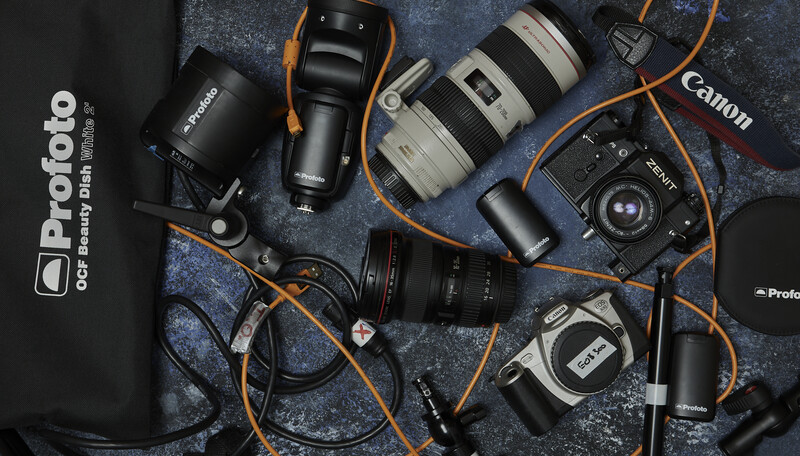 Photography equipment laid out flat including cameras, lenses, lighting gear, and accessories on a textured surface.