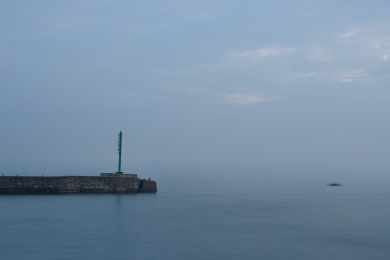 Minimalist seascape with a stone pier and green lighthouse structure in calm blue-grey waters at dusk.