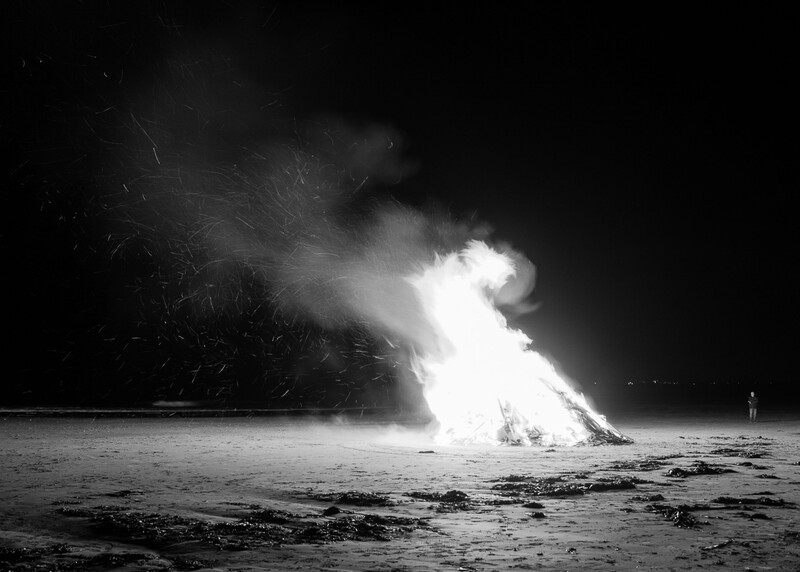 Large bonfire engulfed in bright white flames and smoke against a dark night sky on a beach.