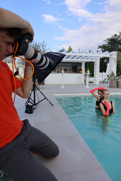 Photographer in red shirt shooting poolside with telephoto lens while subject swims in turquoise water.