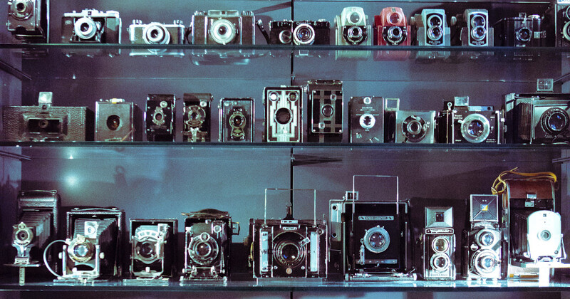 Collection of vintage film cameras displayed on three glass shelves with dramatic blue-tinted lighting.