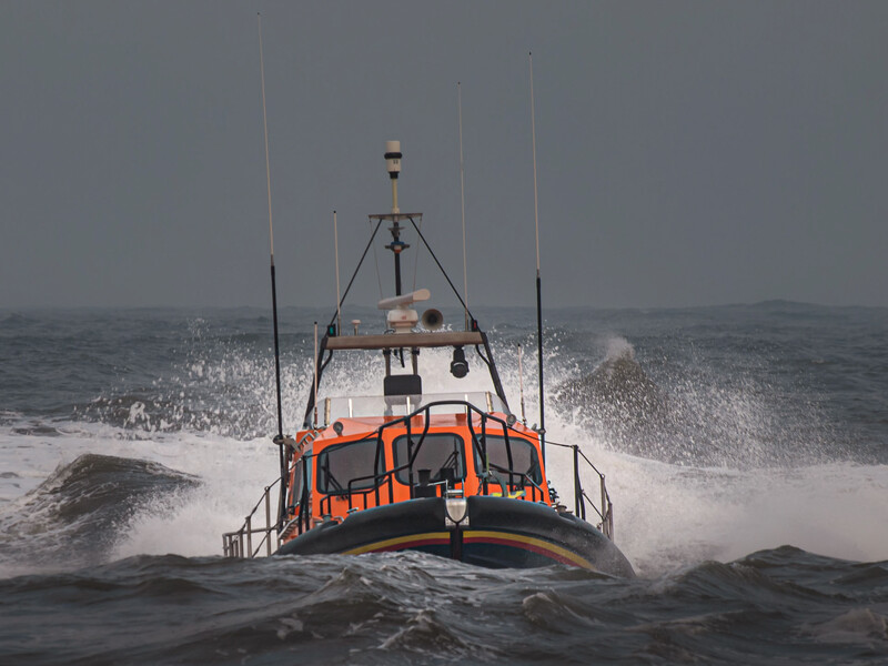 Orange and white lifeboat navigating through rough ocean waves with spray and overcast sky.