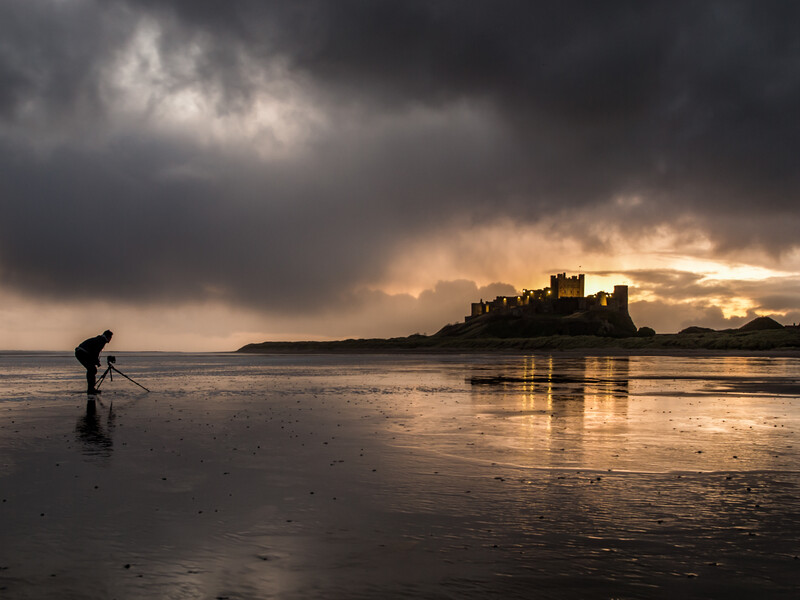 Person wading in shallow water on a beach at sunset, facing a distant castle on an island.