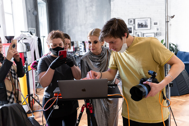 Five people in a studio setting review work on a laptop while holding cameras and phones.