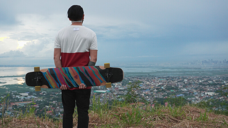 Person holding a colorful striped wakeboard while standing on a hilltop overlooking a sprawling coastal city and bay.