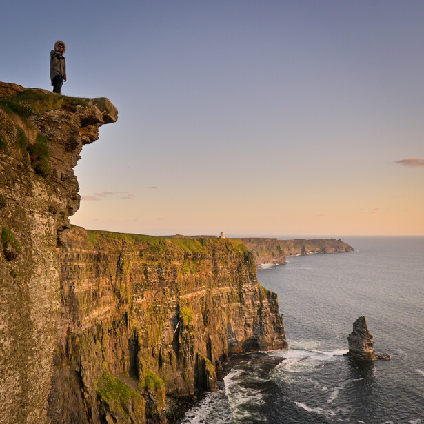 Hiker standing on a rocky cliff overlooking coastal cliffs and sea stacks at golden hour.