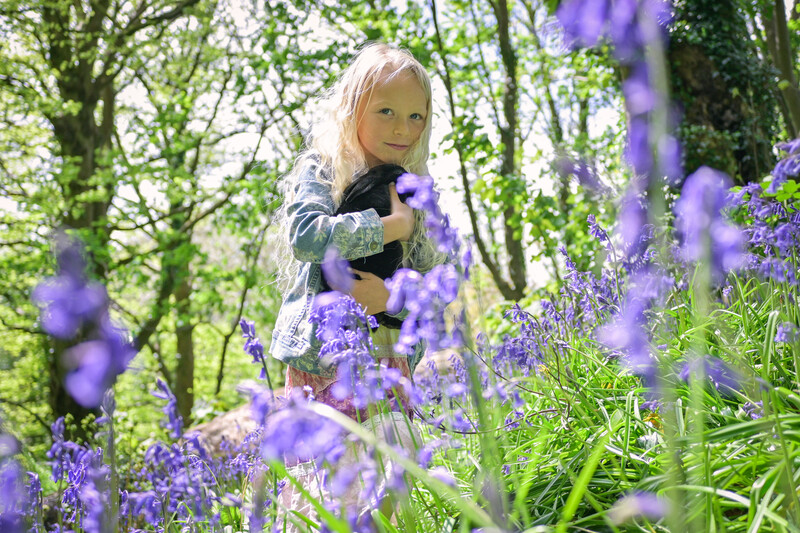 Young child with blonde hair among blooming purple flowers in a woodland setting.