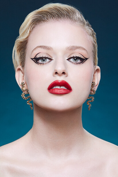 Woman with blonde hair, bold red lipstick, and graphic winged eyeliner against a teal background.