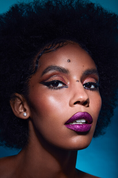 Beauty portrait with bold purple lip color and graphic winged eyeliner against blue background.