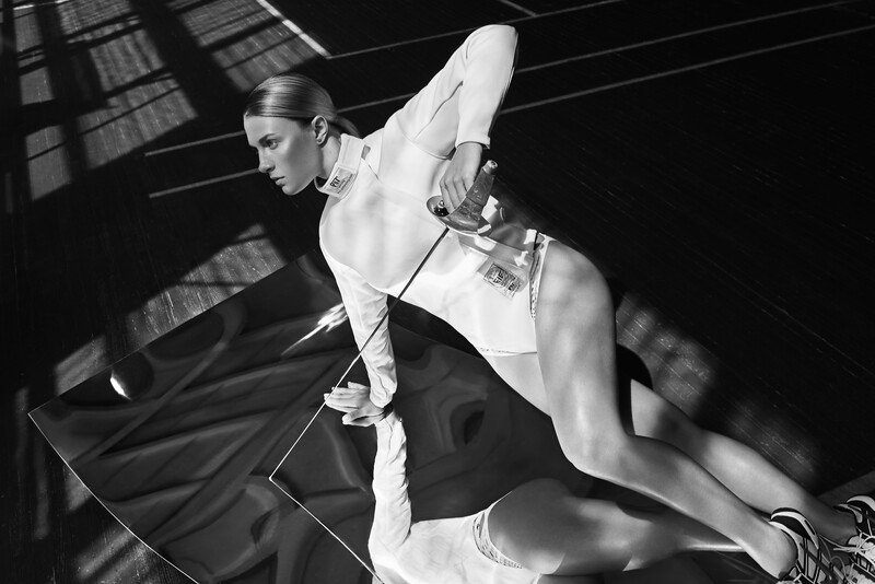 Black and white portrait of a woman in athletic wear posing dynamically with dramatic window light creating linear shadows across her body.