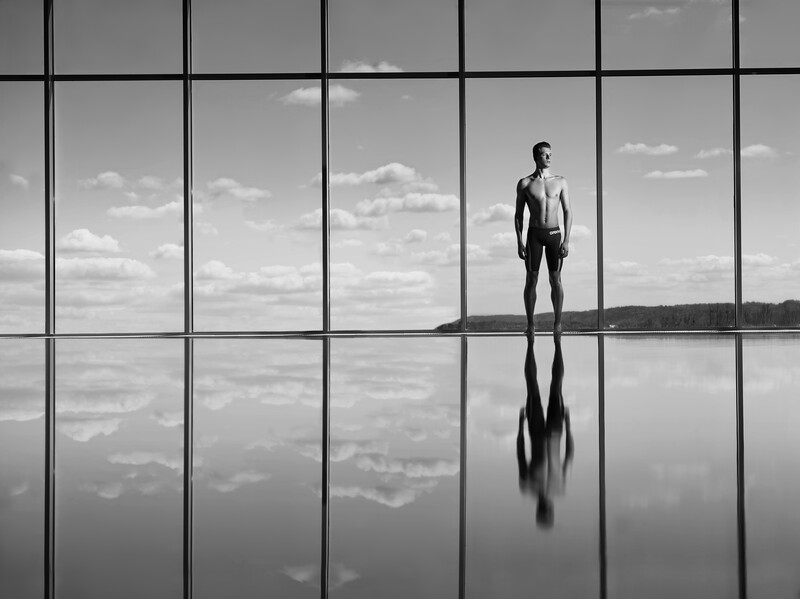 Solitary figure standing on reflective surface with grid-pattern windows and sky backdrop.