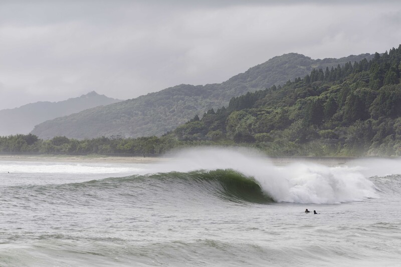 Ocean wave breaking near forested coastline with misty mountains in background.