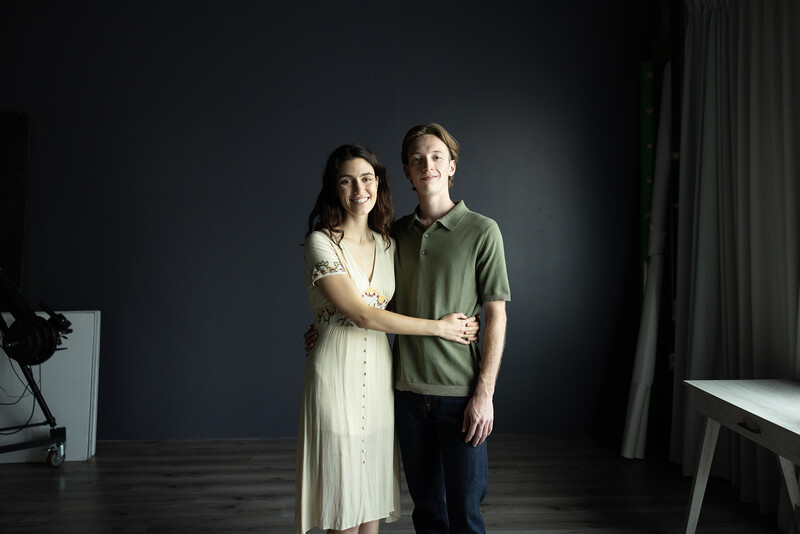 Two people standing together against a dark backdrop, lit by studio lighting.