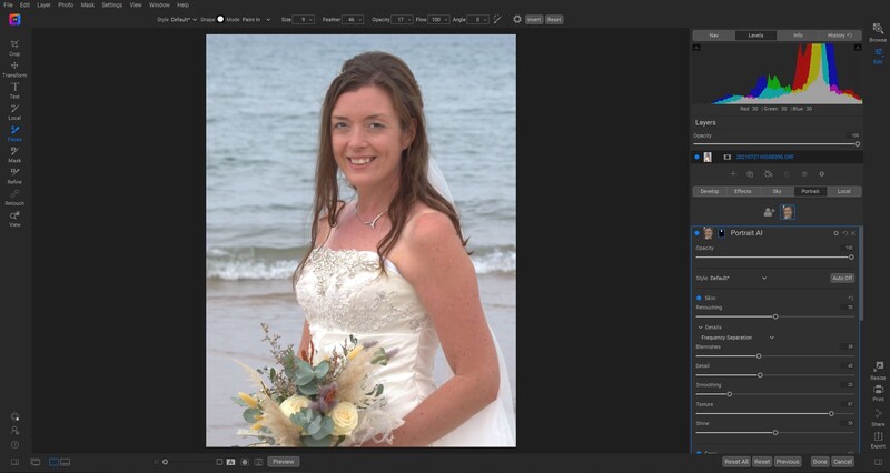 Bride in white beaded strapless gown holding wildflower bouquet on beach with ocean waves behind her.