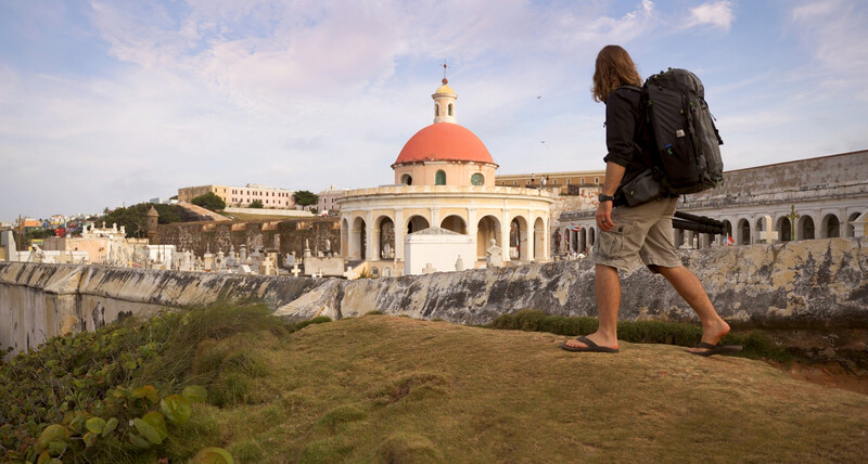 Backpacker with hiking pack standing on grassy overlook facing a domed colonial church and fortification walls.