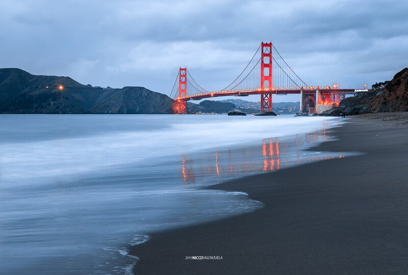 Golden suspension bridge at dusk with long-exposure waves on dark sand beach in foreground.