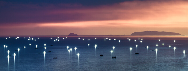 Boats at anchor in calm water under a twilight sky, illuminated by mooring lights reflecting upward.